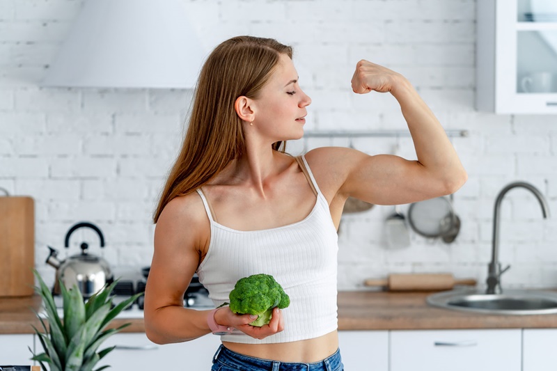 Mulher forte com brócolis na cozinha. Jovem saudável mostra seus braços fortes. Magnus Personal Trainer